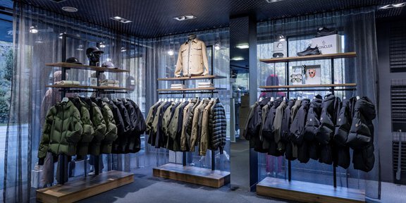 Ein moderner Bekleidungsladen mit verschiedenen Jacken und Mänteln auf Holzregalen. Das elegante Ambiente wird durch eine gedämpfte Beleuchtung und durchsichtige Vorhänge ergänzt. - Sport4You Filiale Avenzio - Sölden
