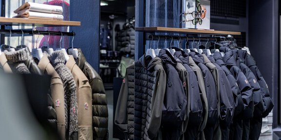 Ein Geschäft mit einer Auswahl an Jacken, die an Kleiderständern hängen. Die Farben sind überwiegend dunkel und die Jacken sind vielfältig in Design und Material. - Sport4You Filiale Avenzio - Sölden