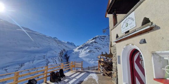 A mountain cabin in a snowy environment with a clear blue sky. In the foreground are backpacks on a wooden terrace. - Hochjoch Hospiz - Vent