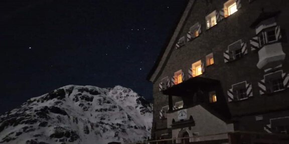 A mountain hut at night, surrounded by snow-covered mountains. Some windows are illuminated and the starry sky is visible. - Hochjoch Hospiz - Vent