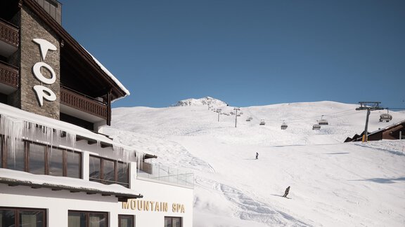 Ein schneebedecktes Berggebiet mit einem Skilift und einer Skipiste. Im Vordergrund steht ein modernes Hotel mit dem Schriftzug "TOP". - TOP Hotel Hochgurgl - Hochgurgl