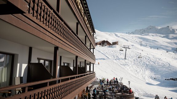Ein wunderschönes Alpengasthaus mit Holzbalustraden und einer schneebedeckten Landschaft im Hintergrund. Skifahrer sind auf den Pisten aktiv, während Gäste auf der Terrasse entspannen. - TOP Hotel Hochgurgl - Hochgurgl