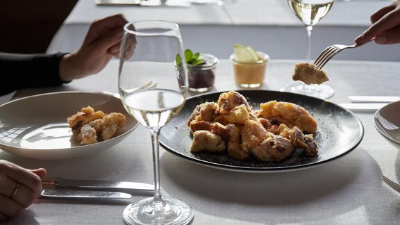 Ein eleganter Tisch mit zwei Gläsern Weißwein und köstlichem frittiertem Essen auf einem schwarzen Teller. Eine Person greift nach einem Stück des Gerichts, während eine andere Portion auf einem zweiten Teller serviert ist. - TOP Hotel Hochgurgl - Hochgurgl