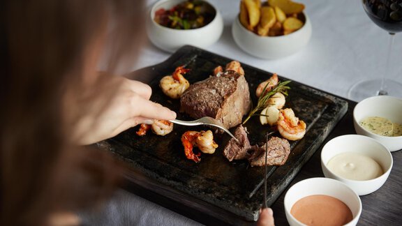 Ein köstliches Steak mit Garnelen auf einem heißen Stein, begleitet von verschiedenen Saucen. Im Hintergrund sind Beilagen in kleinen Schalen zu sehen. - TOP Hotel Hochgurgl - Hochgurgl