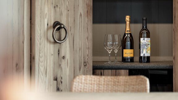 Two bottles of wine are standing on a wooden shelf, next to two glasses. The background is designed simply and modern. - Chalet Cecilia by MYALPS - Obergurgl 
