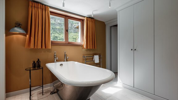 A modern bathroom with a freestanding bathtub and a large window. The walls are painted in a warm orange and the lighting creates an inviting atmosphere. - Chalet Cecilia by MYALPS - Obergurgl 