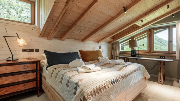 A cozy bedroom with wooden walls and slanted ceilings. It is furnished with a bed, a dresser, and large windows that offer a beautiful view. - Chalet Cecilia by MYALPS - Obergurgl 