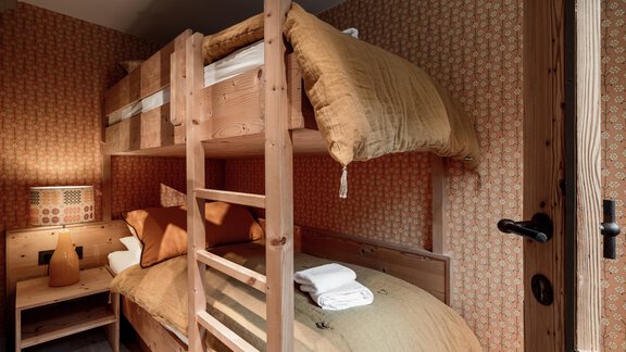 A cozy room with a wooden bunk bed. The walls are decorated with a warm pattern, and there is a small bedside lamp. - Chalet Cecilia by MYALPS - Obergurgl 