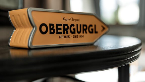 A striking yellow sign indicates the direction to Obergurgl with the distance to Reims. It is placed on a table and has a modern, appealing shape. - Chalet Cecilia by MYALPS - Obergurgl 