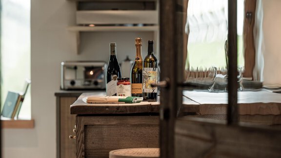 A modern kitchen area with a wooden table and various wine bottles on the countertop. In the background, a microwave and a bright window front are visible. - Chalet Cecilia by MYALPS - Obergurgl 