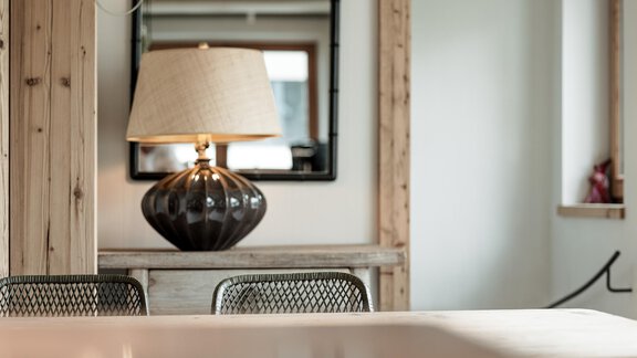 A stylish lamp with a black lampshade stands on a wooden table. In the background, a frame is reflected in the mirror. - Chalet Cecilia by MYALPS - Obergurgl 