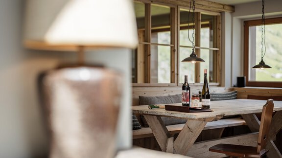 A cozy dining area with a wooden table and two bottles of wine. Soft light from a lamp and large windows create an inviting atmosphere. - Chalet Cecilia by MYALPS - Obergurgl 