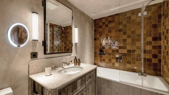 A modern bathroom with a bathtub and a wooden sink. The walls are designed with decorative tiles, and a large mirror provides additional brightness. - Chalet Cecilia by MYALPS - Obergurgl 