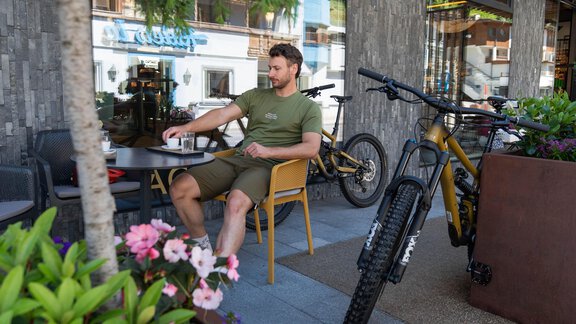 Ein Mann sitzt entspannt an einem Tisch vor einem Café, umgeben von Blumen. Fahrräder sind neben ihm abgestellt, während die Straße im Hintergrund zu sehen ist. - Glanz & Glory Appartement Sölden - Sölden