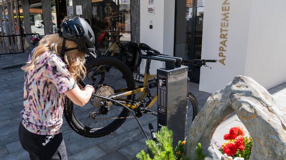Eine Person mit einem Helm repariert ein Fahrrad vor einem modernen Gebäude mit der Beschriftung "APPARTEMENTS". Im Vordergrund blühen bunte Pflanzen. - Glanz & Glory Appartement Sölden - Sölden