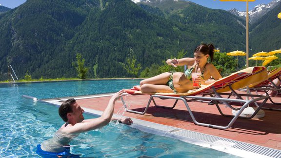 A man is swimming in the pool while a woman is relaxing on a lounger. In the background, green mountains and a clear sky can be seen. - Vivea 4* Hotel Umhausen im Ötztal - Umhausen