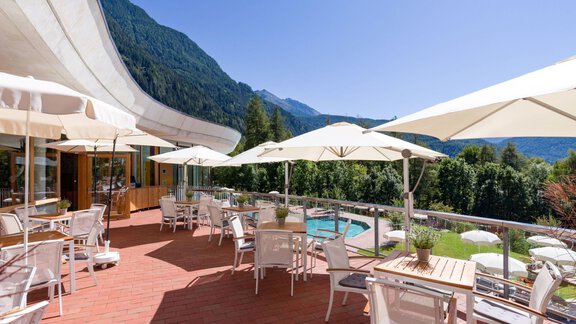 A sunny terrace with white tables and chairs. In the background, mountains and a pool can be seen. - Vivea 4* Hotel Umhausen im Ötztal - Umhausen