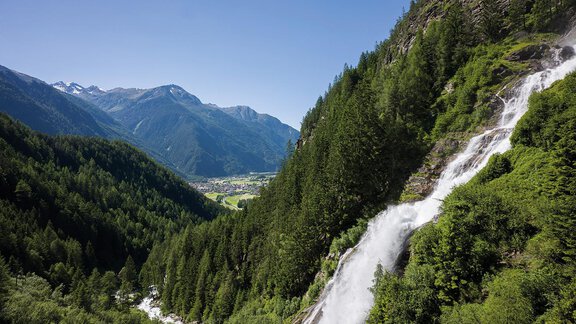 A breathtaking waterfall cascades over green rocks into a green valley. In the background, majestic mountains and a clear blue sky are visible. - Vivea 4* Hotel Umhausen im Ötztal - Umhausen