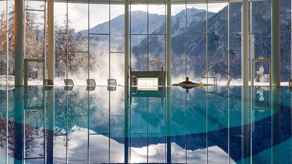 A modern wellness area with a large indoor pool. Through large windows, one looks out at snow-covered mountains and a clear sky. - Vivea 4* Hotel Umhausen im Ötztal - Umhausen