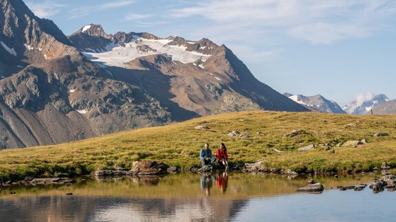 Two people are sitting by the shore of a clear mountain lake in a picturesque landscape. In the background, majestic mountains rise with snow-capped peaks. - Hotel Alt Vent - Tyrol - Vent