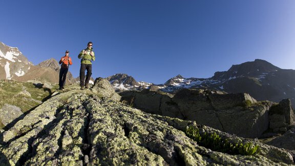 Two hikers are exploring a rocky path in the mountains. The sky is clear and the surroundings are stunning. - Hotel Vent - Vent