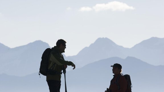 Two hikers stand on a mountainside and are having a conversation. In the background, the silhouette of mountains and a clear sky can be seen. - Hotel Vent - Vent