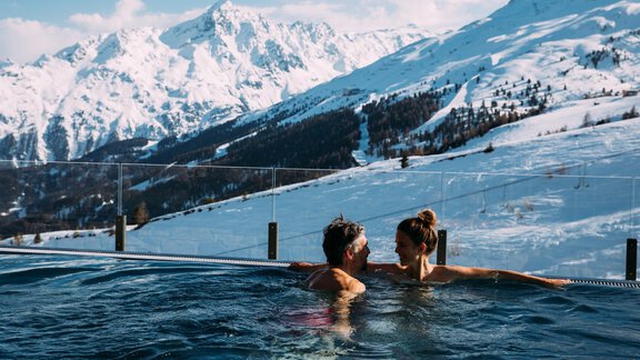 Ein Paar entspannt in einem Außenpool mit schneebedeckten Bergen im Hintergrund. Die Winterlandschaft bietet eine atemberaubende Kulisse für eine romantische Auszeit. - Hotel Enzian - Hochsölden
