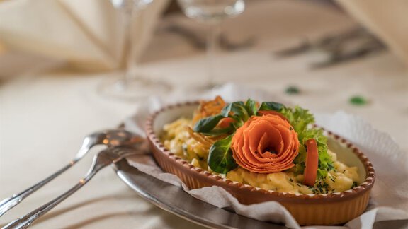 Ein köstliches Gericht in einer keramischen Form, dekoriert mit frischen Kräutern und einer ansprechenden Gemüseblume. Der Tisch ist elegant gedeckt, mit Gläsern und Besteck im Hintergrund. - Hotel Tauferberg - Niederthai