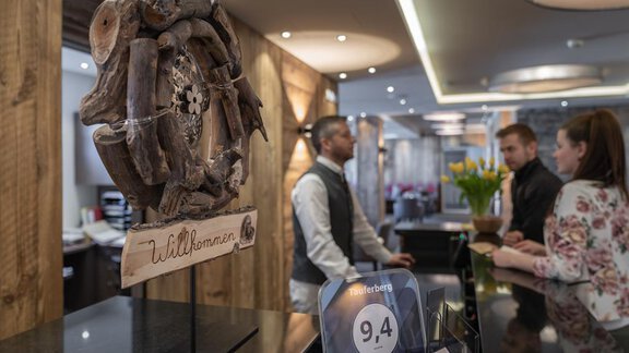 Eine moderne Hotelrezeption mit Holzdekoren und einem Willkommensschild. An der Rezeption stehen Gäste, die eingecheckt werden. - Hotel Tauferberg - Niederthai
