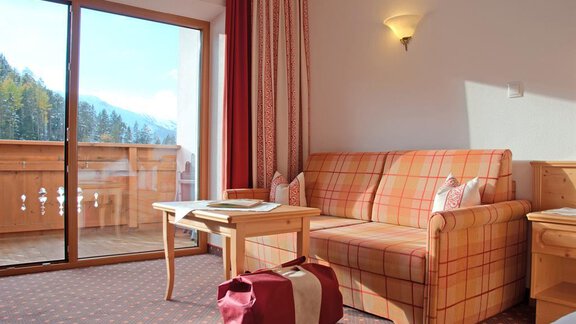 Ein gemütliches Zimmer mit einem karierten Sofa und einem Holztisch. Große Fenster bieten einen Blick auf den Balkon und die umliegende Natur. - Hotel Tauferberg - Niederthai