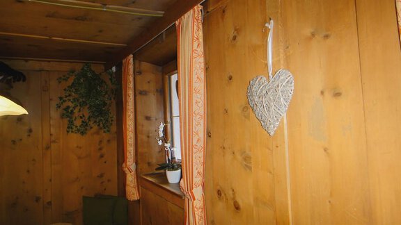A cozy room with wood paneling and a large window opening. A heart-shaped decorative element hangs on the wall, and some plants are visible. - Haus Elisabeth - Vent