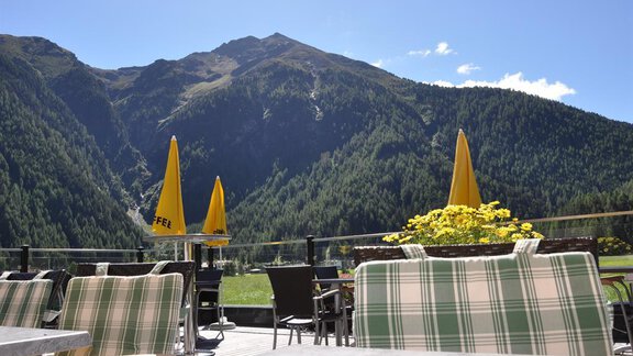 Eine gemütliche Terrasse mit Stühlen und Sonnenschirmen, die auf eine beeindruckende Berglandschaft blickt. Grüne Wälder und blauer Himmel schaffen eine einladende Atmosphäre. - Hotel Tauferberg - Niederthai