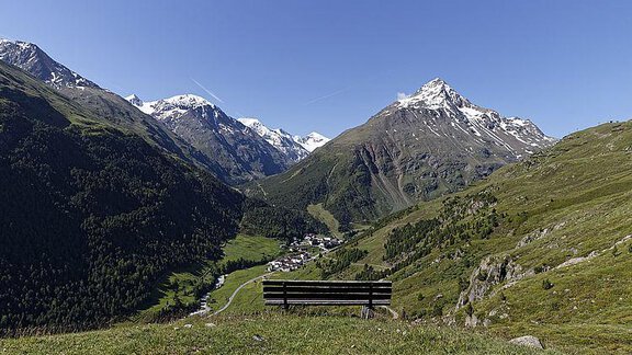 A picturesque mountain landscape with snow-covered peaks and green meadows. In the foreground stands an empty bench overlooking the valley and the village. - Hotel Alt Vent - Tyrol - Vent