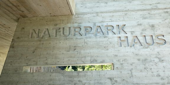 A modern building with the inscription "NATURE PARK HOUSE" on a concrete wall. There is a window that offers views of nature. - Naturpark Ötztal - Naturpark Haus - Längenfeld