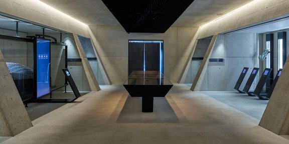 Ein moderner Raum mit Betonwänden und schlichten, geometrischen Formen. In der Mitte befindet sich ein schwarzer Tisch, umgeben von digitalen Displays und Maschinen. - 007 ELEMENTS - Sölden