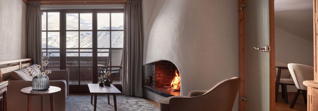 Ein gemütlicher Wohnraum mit Holzdecke und großen Fenstern, die Blick auf die Berge bieten. In der Mitte steht ein Tisch mit bequemen Stühlen und einem Kamin, der für Wärme sorgt. - TOP Hotel Hochgurgl - Hochgurgl