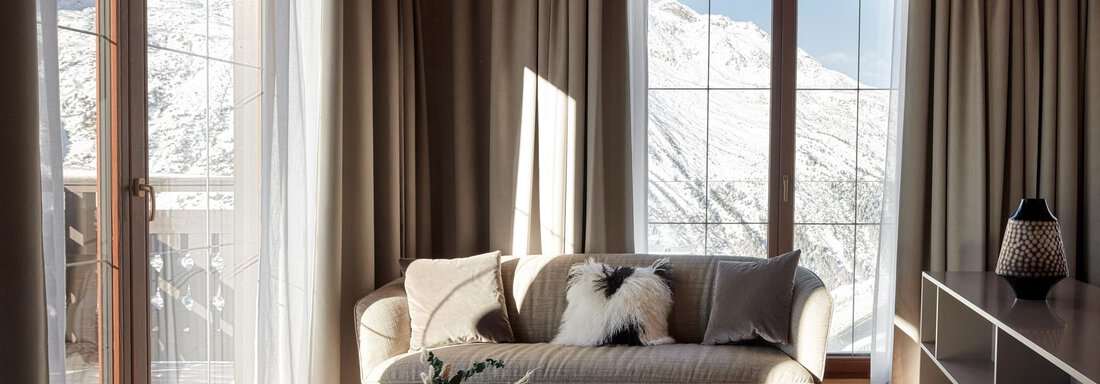 Ein modernes Wohnzimmer mit großen Fenstern, die auf verschneite Berge blicken. Die Einrichtung ist hell und gemütlich mit einem Sofa und einem eleganten Couchtisch. - TOP Hotel Hochgurgl - Hochgurgl