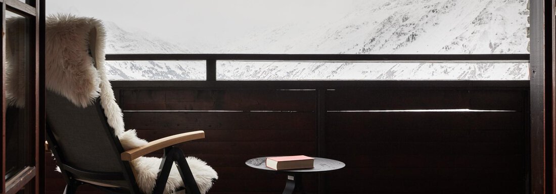 Ein gemütlicher Balkon mit einem bequemen Stuhl und einem kleinen Tisch. Im Hintergrund sind Schneebedeckte Berge unter einem grauen Himmel zu sehen. - TOP Hotel Hochgurgl - Hochgurgl