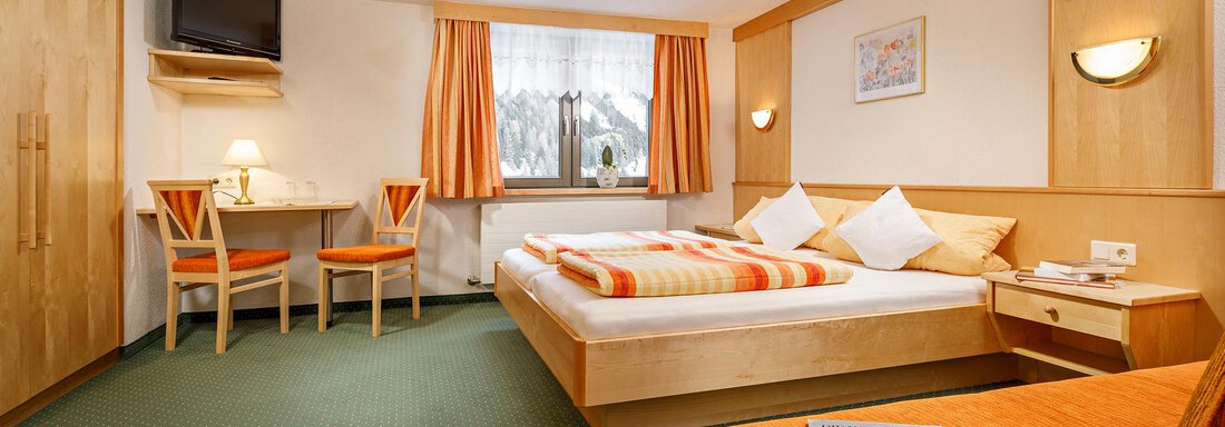Ein gemütliches Zimmer mit einem großen Bett und orangefarbenen Akzenten. Es gibt einen Tisch, einen Fernseher und Fenster mit Vorhängen. - Hotel Tauferberg - Niederthai