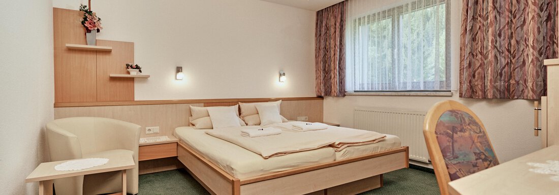 Ein gemütliches Schlafzimmer mit einem Doppelbett und grünem Teppich. Neben dem Bett stehen ein Tisch und ein Stuhl sowie ein Fenster mit Vorhängen. - Hotel Tauferberg - Niederthai
