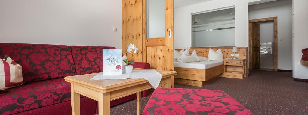 Ein gemütliches Zimmer mit rotem Sofa und Hocker. Das Schlafzimmer ist hell und freundlich mit einem Doppelbett und Holzregalen. - Hotel Tauferberg - Niederthai