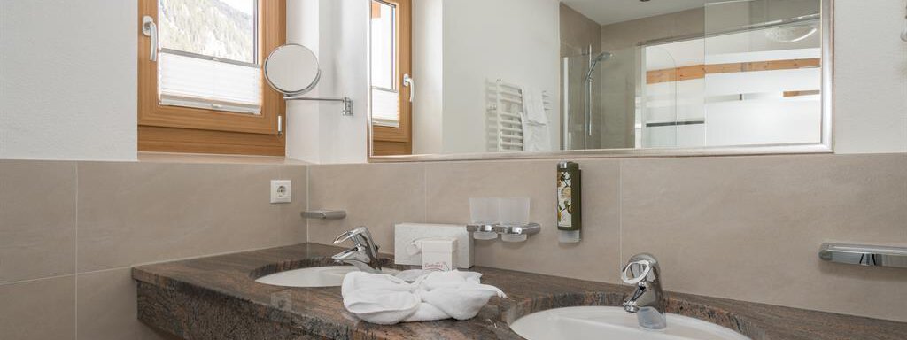 Ein modernes Badezimmer mit zwei Waschbecken und einem großen Spiegel. Die Räume sind hell und gut beleuchtet mit Fenstern, die Tageslicht hereinlassen. - Hotel Tauferberg - Niederthai