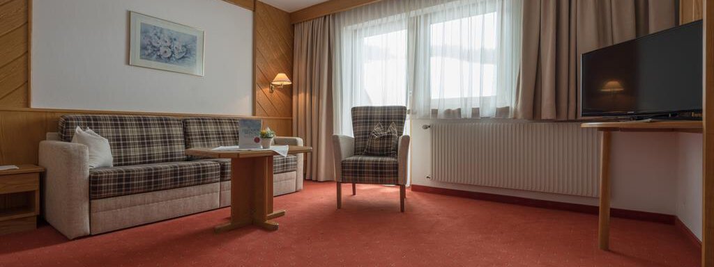 Ein gemütliches Wohnzimmer mit einem Sofa, einem Sessel und einem Tisch. Große Fenster lassen viel Licht herein, und der Teppich ist rot. - Hotel Tauferberg - Niederthai