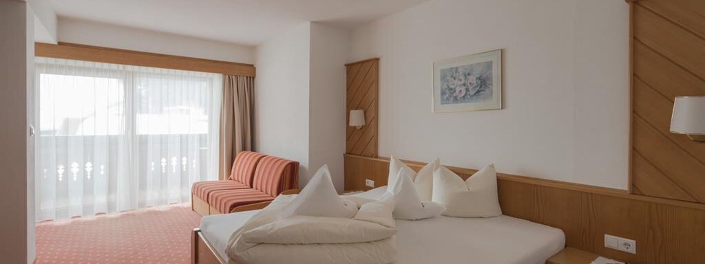 Ein helles Zimmer mit einem großen Bett und dekorativen Kissen. Es gibt ein Sofa und große Fenster, die viel Tageslicht hereinlassen. - Hotel Tauferberg - Niederthai