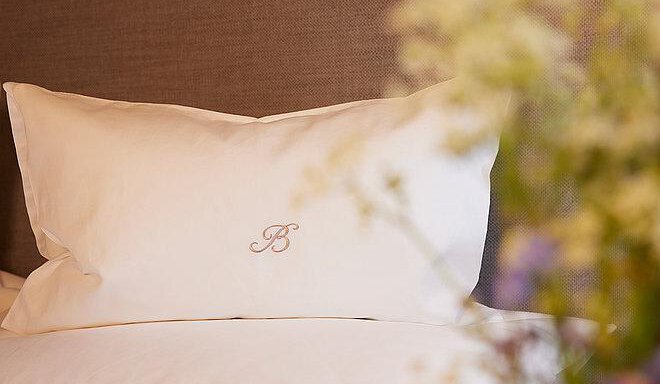 An elegant pillow with a subtle, embroidered initial "B". In the background, soft flowers are visible, creating an inviting atmosphere. - Art & Relax Hotel Bergwelt - Obergurgl 