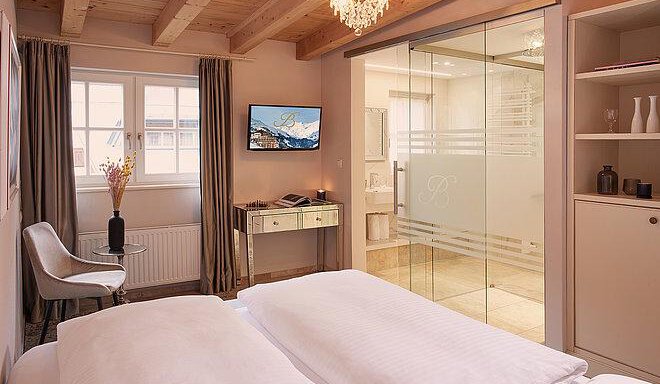 A modern bedroom with a large bed and bright decor.  

Large windows let in plenty of light, and the bathroom is separated by a glass door. - Art & Relax Hotel Bergwelt - Obergurgl 