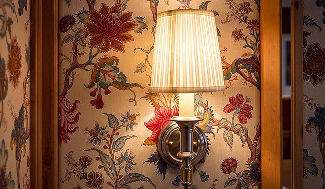 An elegant wall lamp with a striped lampshade. In the background, there is a floral wallpaper with vibrant colors. - Art & Relax Hotel Bergwelt - Obergurgl 