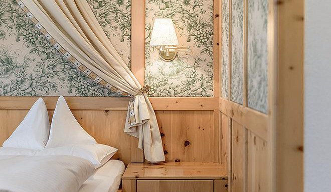 A cozy bedroom with wooden panels and a floral wallpaper. A bedside lamp provides soft lighting. - Art & Relax Hotel Bergwelt - Obergurgl 