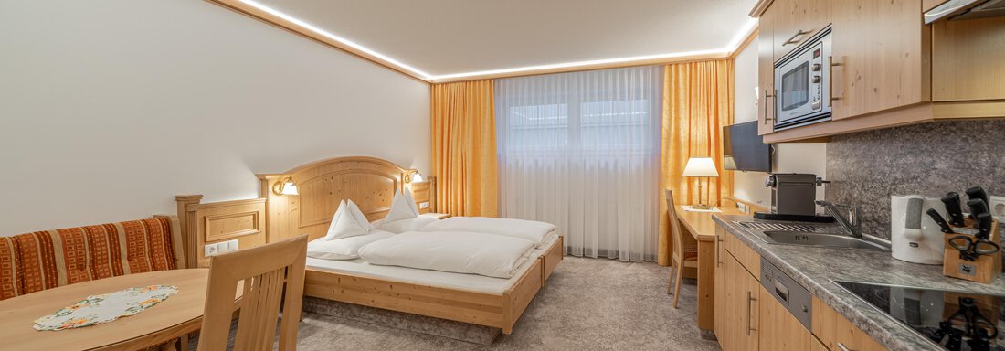 Wohn-Schlafraum A bright hotel room with a double bed, a table, and chairs. The modern kitchenette is well-equipped and the room has large windows with curtains. - Ferienwohnung de Luxe Alpenresidenz am Mühlbach - Obergurgl