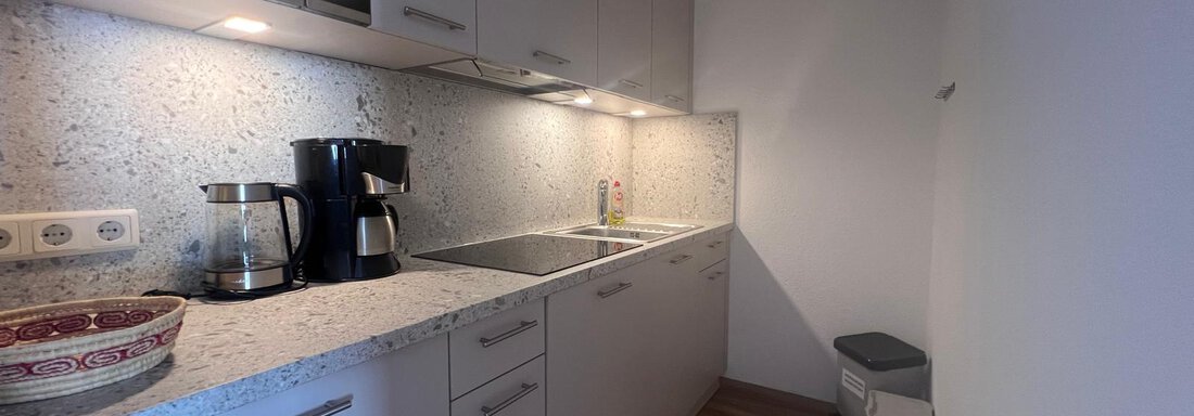 A modern kitchen with a bright granite countertop and simple white cabinets. There is a coffee machine, a faucet, and a trash can in the corner. - Ferienwohnung Fabian - Obergurgl 